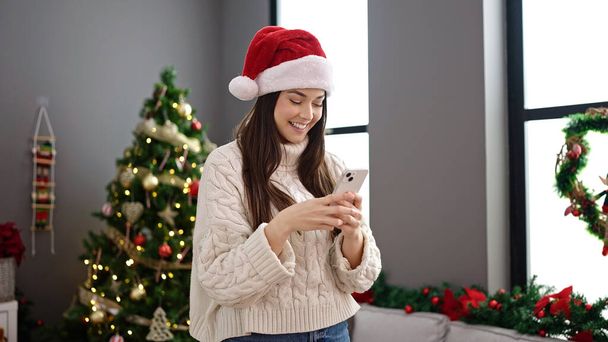 Joven mujer hispana hermosa usando teléfono inteligente de pie junto al árbol de Navidad en casa - Foto, imagen