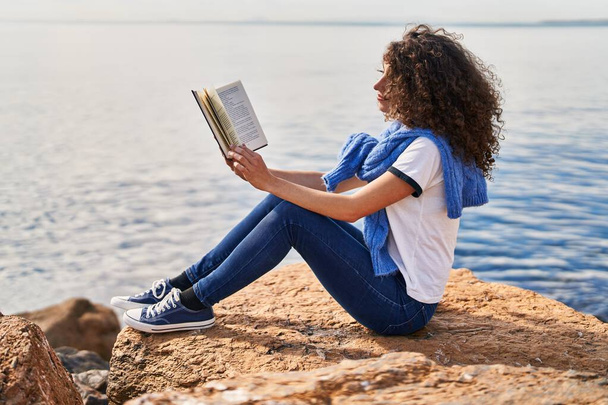 Young hispanic woman reading book sitting on rock at seaside - Foto, imagen
