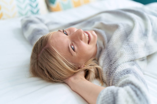 Joven mujer rubia sonriendo confiada acostada en la cama en el dormitorio - Foto, imagen