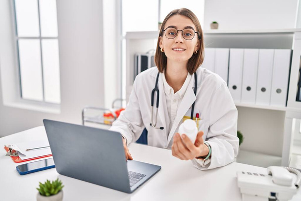 Young caucasian woman doctor using laptop holding pills at clinic - Foto, afbeelding