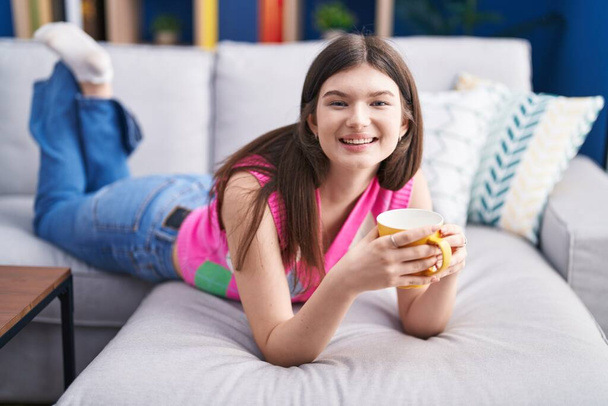 Junge Kaukasierin trinkt Kaffee liegend auf Sofa zu Hause - Foto, Bild