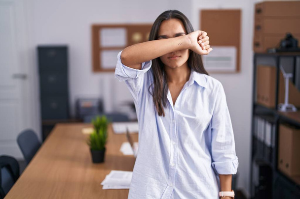 Młoda latynoska kobieta w biurze zasłania oczy ramieniem, wygląda poważnie i smutno. Koncepcja niewidoczności, ukrywania i odrzucania  - Zdjęcie, obraz