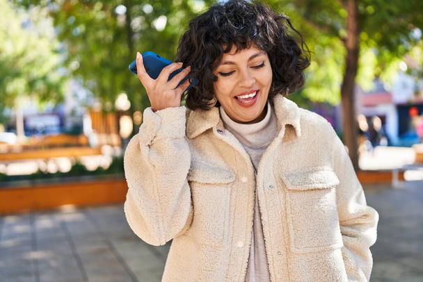 Молодая красивая латиноамериканка улыбается уверенно слушая аудиосообщение от смартфона в парке - Фото, изображение