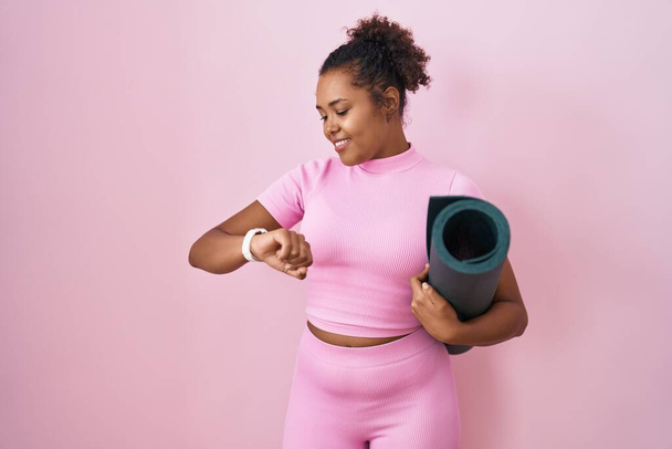 Junge hispanische Frau mit lockigem Haar hält Yogamatte über rosa Hintergrund und überprüft die Uhrzeit an der Armbanduhr, entspannt und selbstbewusst  - Foto, Bild
