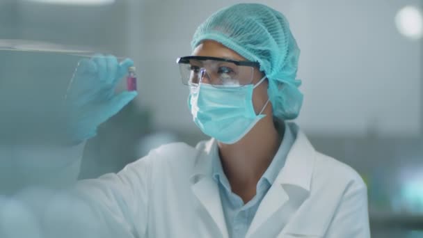Química asiática en mascarilla protectora, sombrero médico y guantes examinando vial con producto químico durante la investigación de laboratorio - Metraje, vídeo
