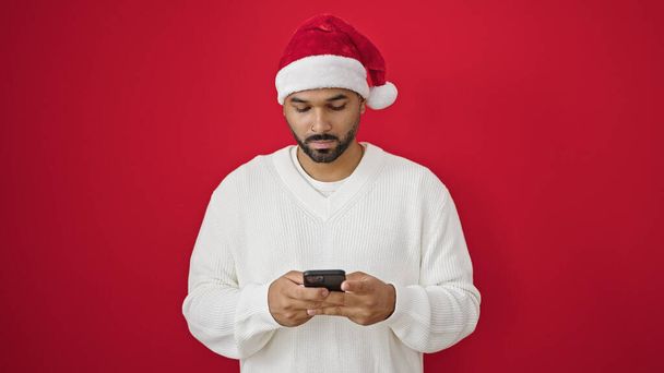 アフリカ系アメリカ人男性は,隔離された赤い背景の上にスマートフォンを使用してクリスマスの帽子をかぶっています - 写真・画像