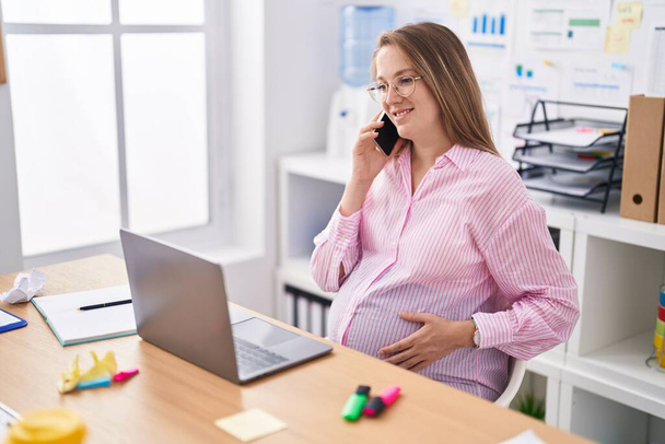 Jeune femme enceinte travailleuse d'entreprise utilisant un ordinateur portable parlant sur smartphone au bureau - Photo, image