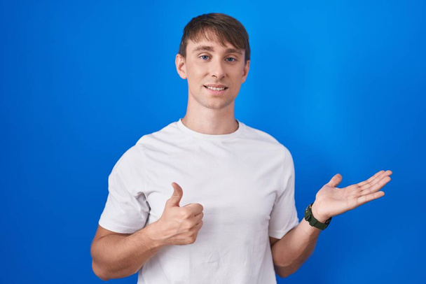 Caucasian blond man standing over blue background showing palm hand and doing ok gesture with thumbs up, smiling happy and cheerful  - Photo, Image