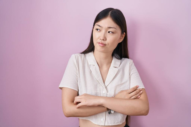 Kínai fiatal nő áll át rózsaszín háttér néz oldalra karok keresztbe tett meggyőződéssel és magabiztossággal  - Fotó, kép