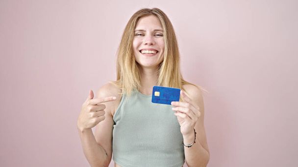 Giovane donna bionda sorridente fiducioso puntando alla carta di credito su isolato sfondo rosa - Foto, immagini