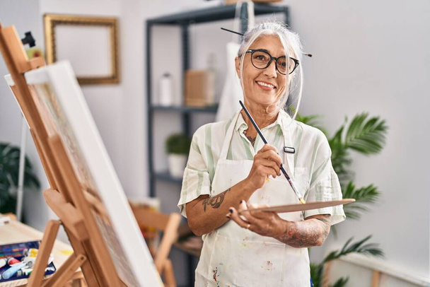 Mujer de pelo gris de mediana edad artista sonriente dibujo seguro en el estudio de arte - Foto, imagen