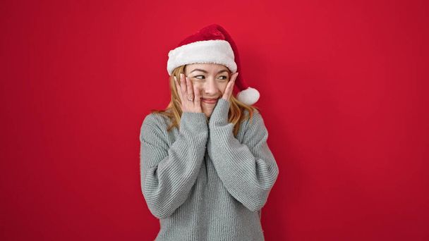 Junge blonde Frau mit Weihnachtsmütze tanzt mit überraschendem Gesichtsausdruck über isoliertem rotem Hintergrund - Foto, Bild