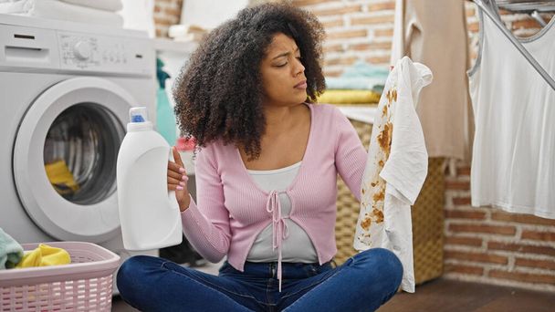 Afričanky americká žena drží saponát láhev a špinavé tričko v prádelně - Fotografie, Obrázek