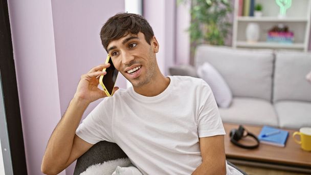 Handsome young hispanic man, oozing confidence, smiling joyfully while engrossed in a phone conversation at home, sitting on his cozy living room sofa, enjoying the positive vibes. - Photo, Image