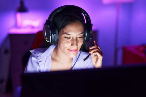 Young beautiful hispanic woman streamer smiling confident sitting on table at gaming room - 写真・画像