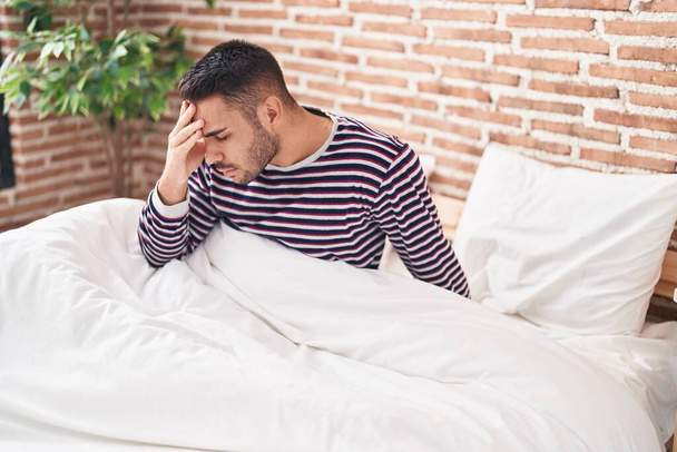 Jonge Spaanse man gestrest zittend op bed in de slaapkamer - Foto, afbeelding