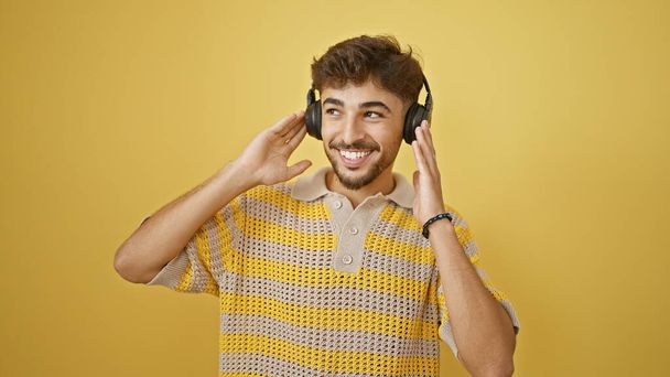 Havalı, kendinden emin genç Arap adam kulaklıklarıyla müzik dinliyor. İzole edilmiş sarı bir duvarın önünde tek başına mutluluk ve mutluluk saçan mutlu bir dans.. - Fotoğraf, Görsel