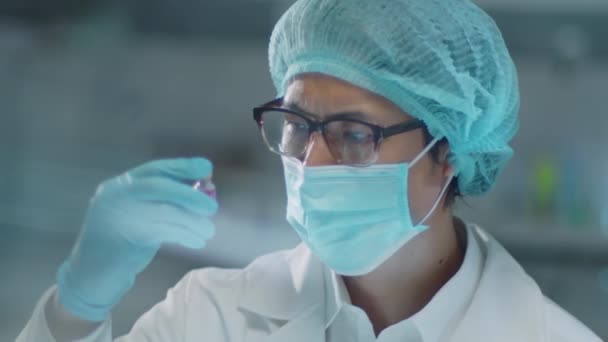 Chest up shot of Asian male chemist in protective face mask, medical hat and gloves examine purple chemical in vial while working in laboratory - Metraje, vídeo