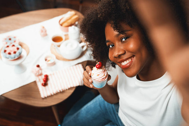 Beautiful young African blogger presenting piece of cupcake in concept special cuisine with selfie on smartphone. Content creating of social media with favorite sweets bakery dish. Tastemaker. - Photo, Image
