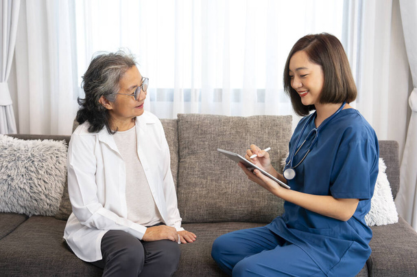 Üniformalı genç Asyalı kadın hemşire evde kıdemli bir hastayı ziyaret ediyor, veri topluyor ve tablet, yaşlanma bakımı, evde sağlık hizmetleri gibi konularda tavsiye veriyor. - Fotoğraf, Görsel