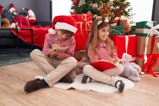 Testvérek rajzolnak füzetbe otthon, karácsonyfa mellett ülve a padlón. - Fotó, kép