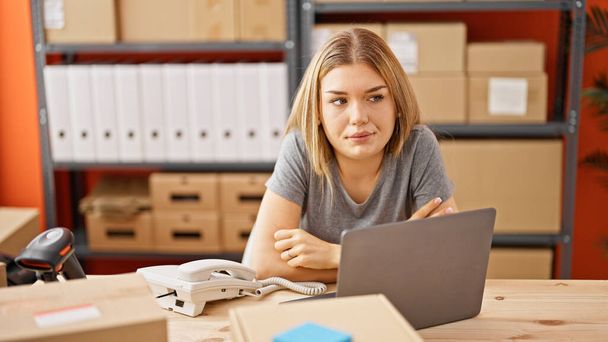 Mujer rubia joven comercio electrónico trabajador de negocios sentado en la mesa con la cara seria en la oficina - Foto, imagen