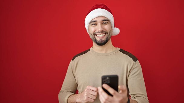 Junger arabischer Mann trägt Weihnachtsmütze mit Smartphone vor isoliertem roten Hintergrund - Foto, Bild