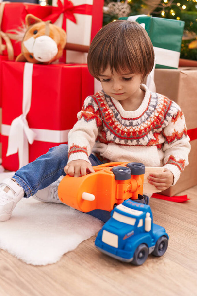 Entzückendes Kleinkind spielt mit LKW-Spielzeug, das zu Hause am Weihnachtsbaum auf dem Boden sitzt - Foto, Bild
