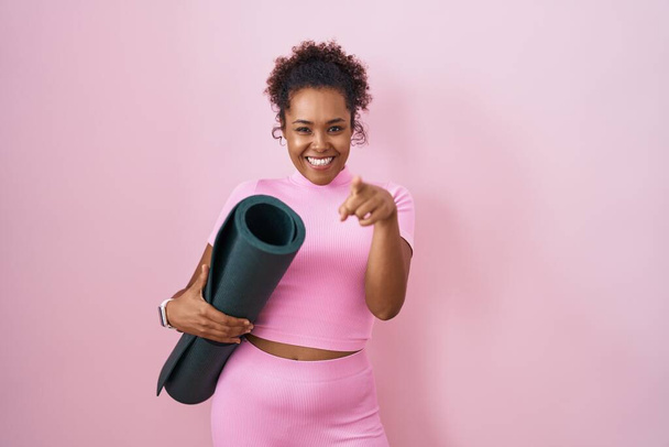 Junge hispanische Frau mit lockigem Haar hält Yogamatte über rosa Hintergrund und zeigt auf Sie und die Kamera mit den Fingern, lächelt positiv und fröhlich  - Foto, Bild