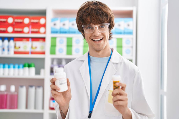 Jovem loiro farmacêutico sorrindo confiante segurando pílulas garrafas na farmácia - Foto, Imagem