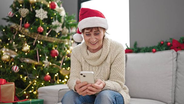 Зрелая латиноамериканка с помощью смартфона сидит у рождественской елки дома - Фото, изображение