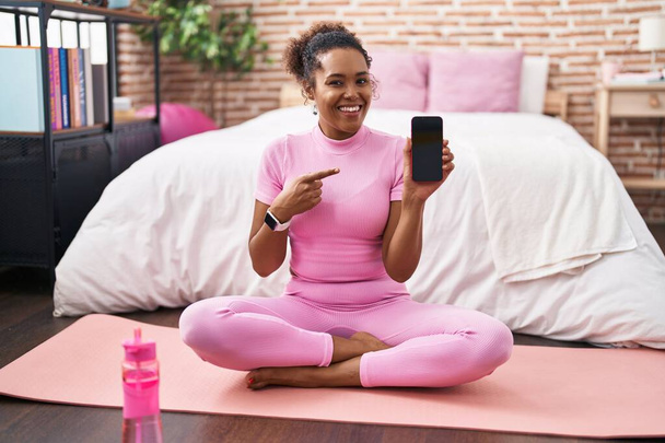 Young hispanic woman with curly hair sitting on yoga mat at the bedroom smiling happy pointing with hand and finger  - Photo, Image