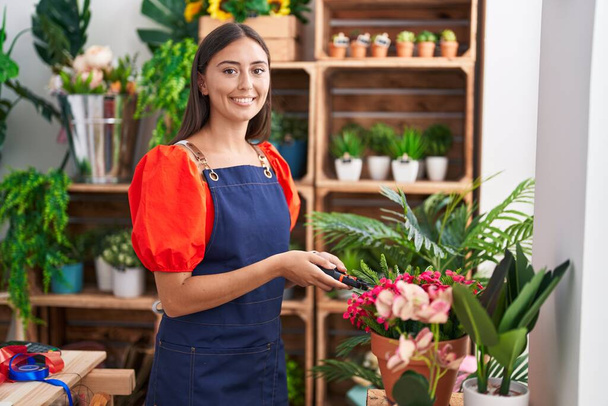 Jovem bela mulher hispânica florista sorrindo confiante folha de corte planta na florista - Foto, Imagem