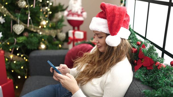 Young beautiful hispanic woman using smartphone and credit card sitting on sofa by christmas tree at home - Photo, Image