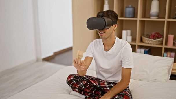 Mosolygó fiatal spanyol játékos srác élvezi ül pizsamában a hálószoba ágy, videojáték segítségével futurisztikus vr szemüveg - Fotó, kép