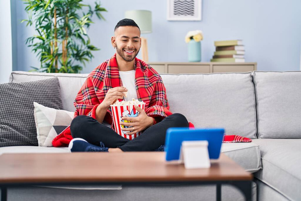 Giovane uomo ispanico mangiare popcorn guardare film su tablet dispositivo sorridente con un sorriso felice e fresco sul viso. mostrando i denti.  - Foto, immagini
