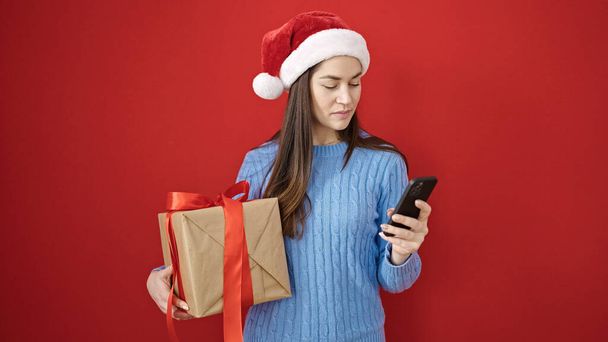 Junge kaukasische Frau trägt Weihnachtsmütze mit Smartphone, das Geschenk über isoliertem roten Hintergrund hält - Foto, Bild