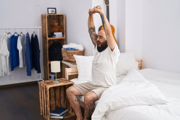Young bald man waking up stretching arms yawning at bedroom - Photo, Image