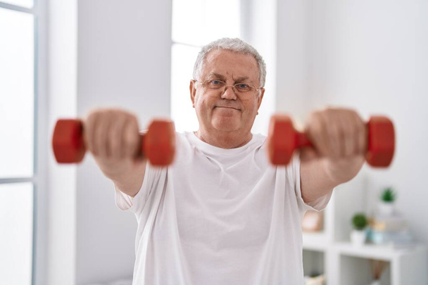 Średniowłosy mężczyzna w średnim wieku uśmiecha się pewnie używając hantli treningowych w domu - Zdjęcie, obraz