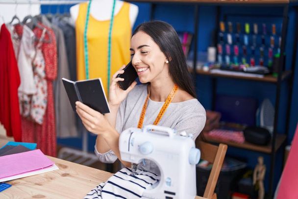 Joven hermosa mujer hispana sastre hablando en el teléfono inteligente portátil de lectura en la fábrica de ropa - Foto, imagen
