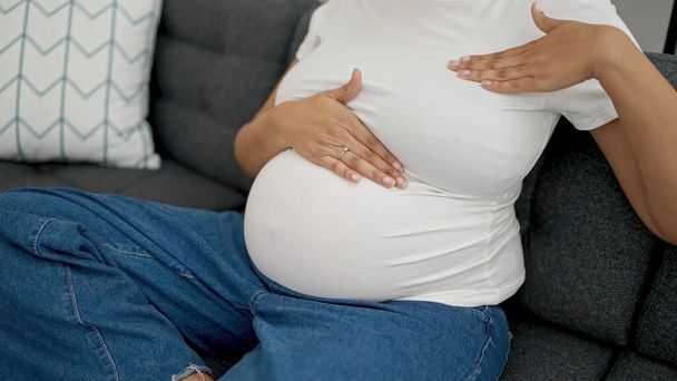 Young pregnant woman examining breast sitting on sofa at home - Valokuva, kuva