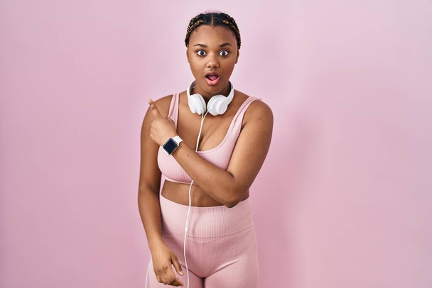 African american woman with braids wearing sportswear and headphones surprised pointing with finger to the side, open mouth amazed expression.  - Photo, Image