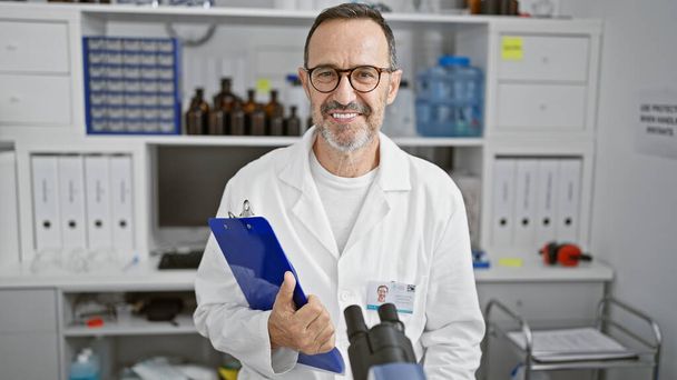 Happy middle age man with grey hair, a confident scientist, joyfully working in his lab with a clipboard - Photo, Image