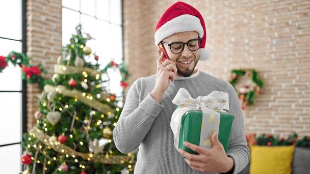 Homme hispanique portant un chapeau de Noël tenant un cadeau parlant au téléphone à la maison - Photo, image
