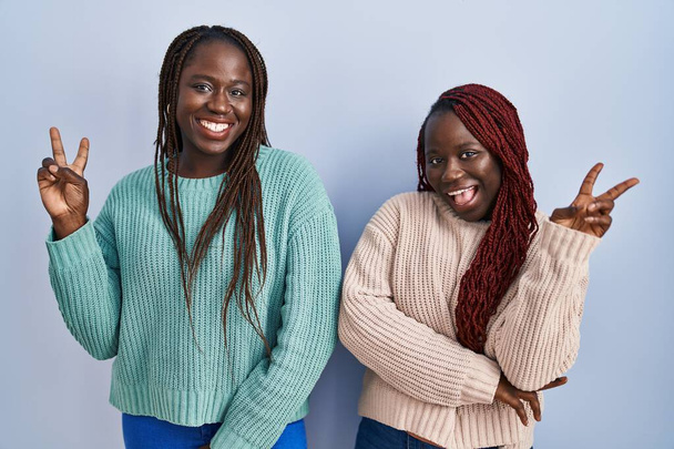 Dvě africké ženy, stojící nad modrým pozadím a usmívající se s veselým obličejem, mrkající na kameru a ukazující prsty na znamení vítězství. číslo dvě.  - Fotografie, Obrázek