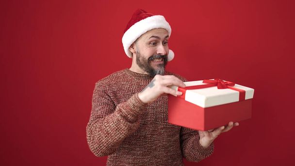 Junger Mann mit Glatze trägt Weihnachtsmütze beim Auspacken von Geschenk vor isoliertem roten Hintergrund - Foto, Bild