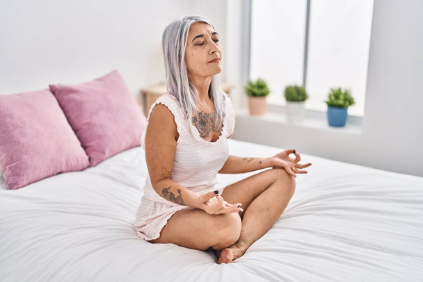 Middelbare leeftijd grijs-harige vrouw doet yoga oefening zittend op bed in de slaapkamer - Foto, afbeelding