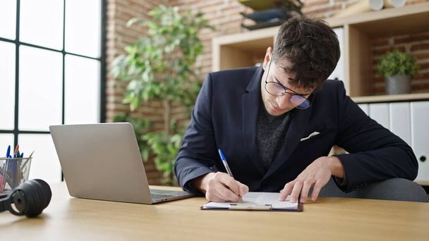 Junger hispanischer Geschäftsmann mit Laptop-Schrift auf Klemmbrett im Büro - Foto, Bild