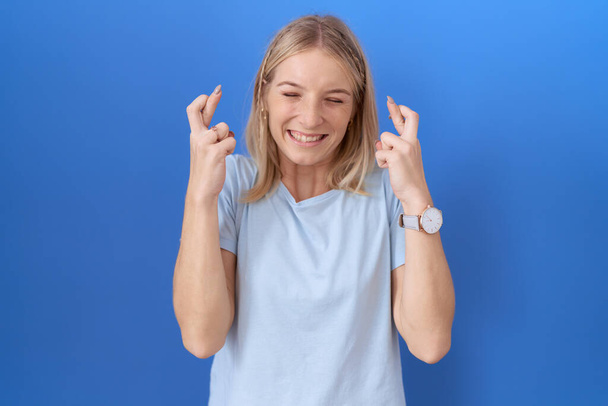Fiatal kaukázusi nő alkalmi kék pólóban gesztikuláló ujjal keresztbe tett mosollyal és csukott szemmel. szerencse és babonás fogalom.  - Fotó, kép