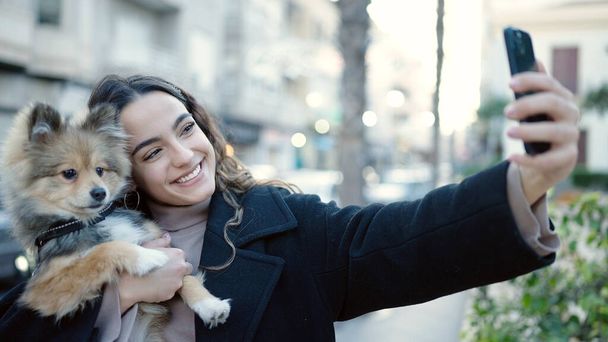 Mladá hispánská žena se psem usměvavý sebevědomí, aby selfie smartphone na ulici - Fotografie, Obrázek